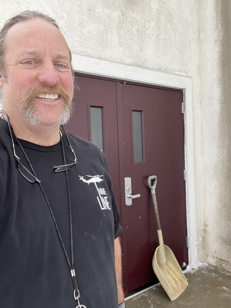 A man smiles in front of a door with a snow shovel leaned against it.