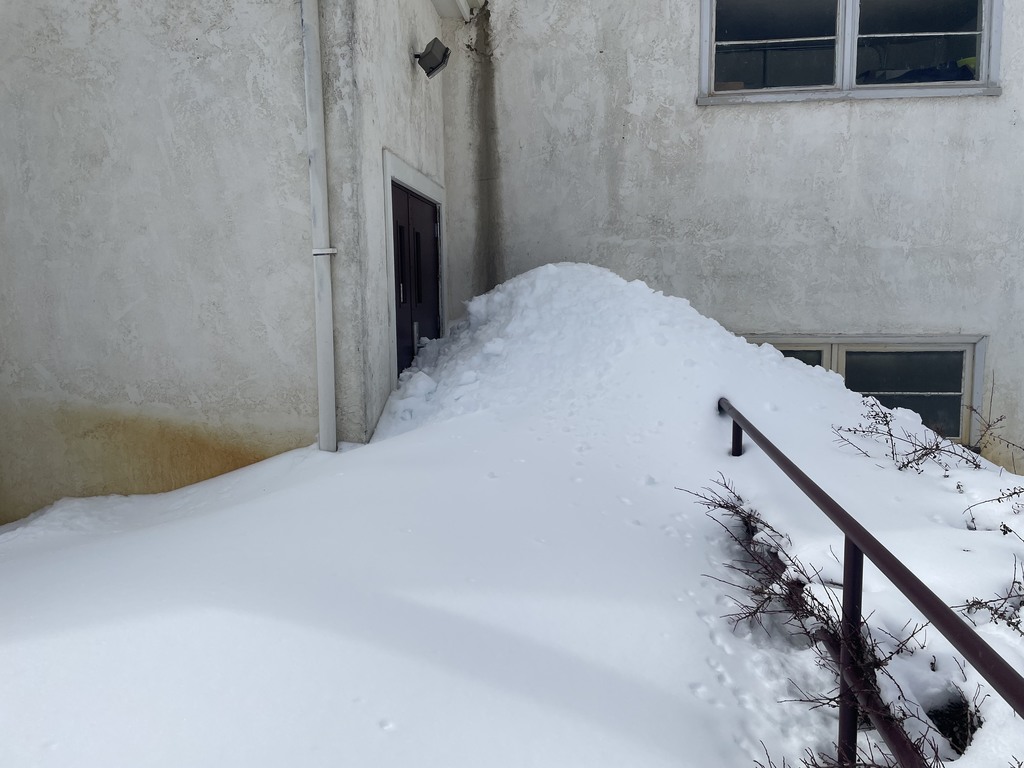 Snow is piled so high, the door is blocked. The hand rail disappears into the snow bank.